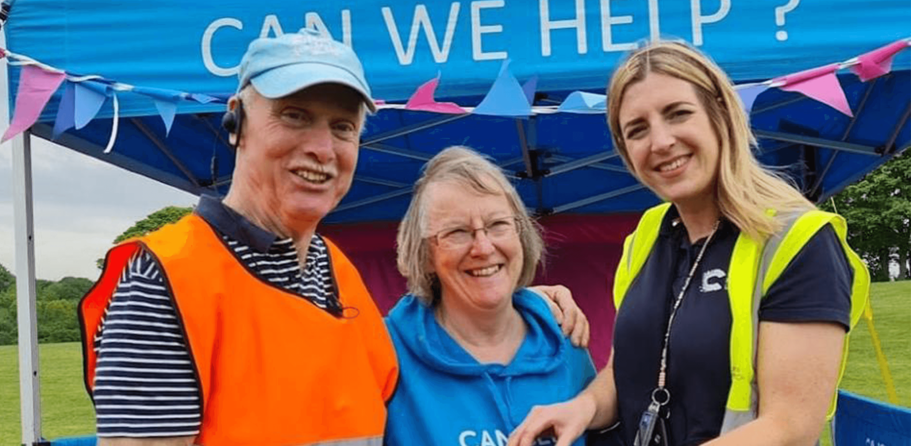 Two Event Volunteers, Phil and Karen, are standing with the Event Manager in front of an enquiries tent. They're all smiling.