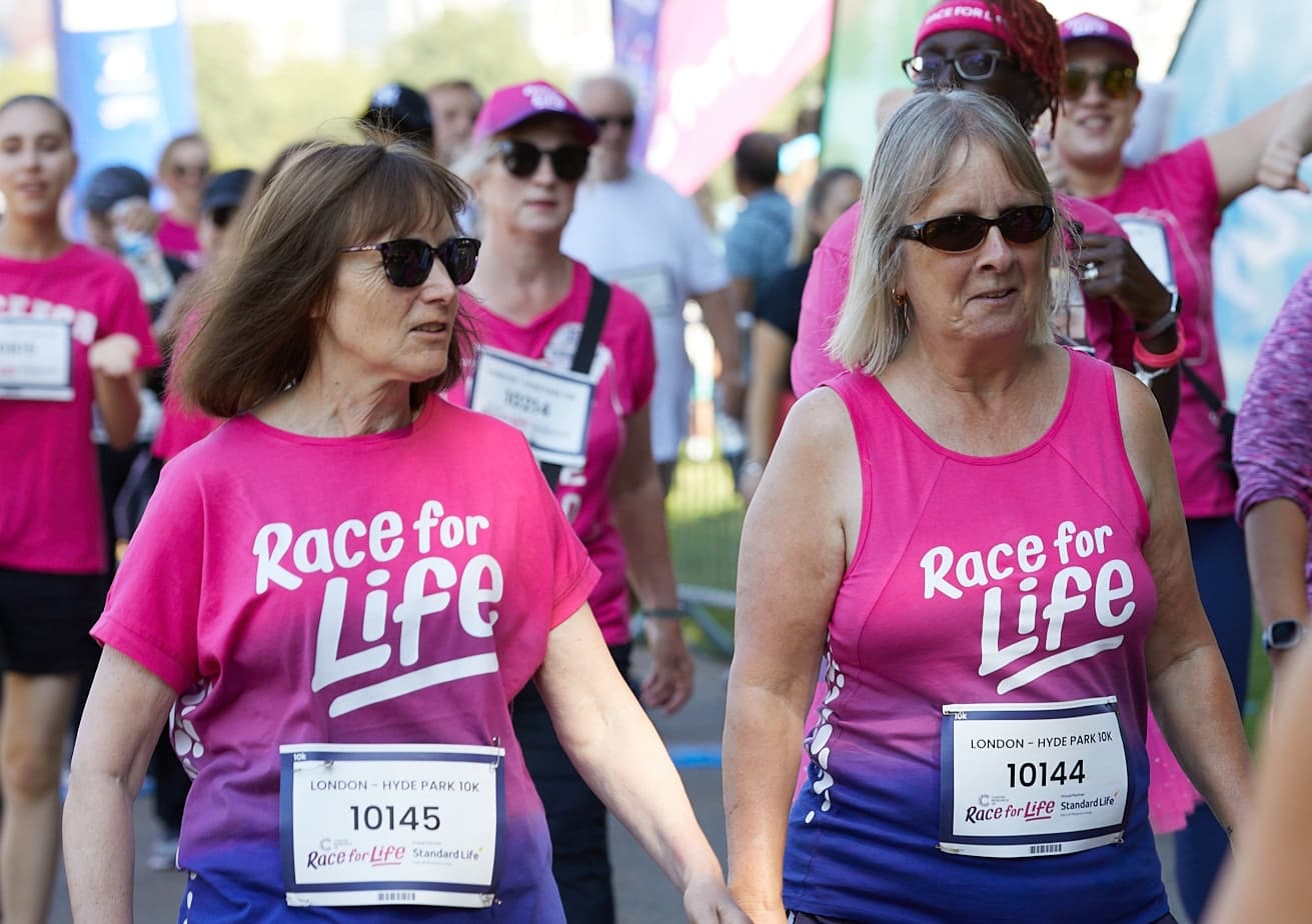Two women wearing pink and purple ombre race for life t-shirts and runners numbers. Both are wearing black sunglasses.